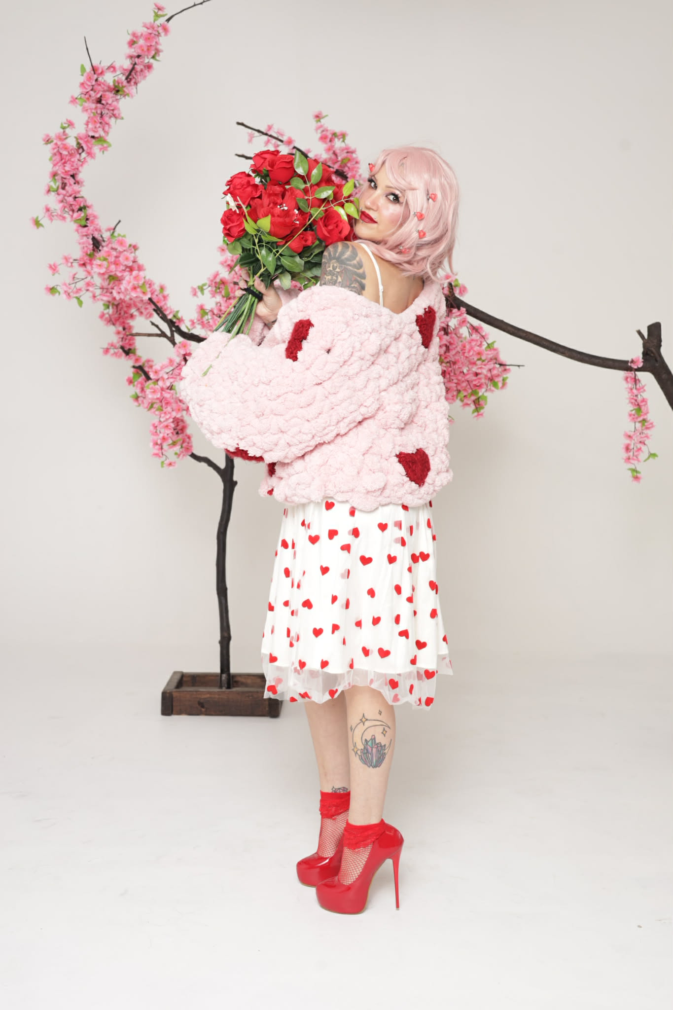 Person with pink hair holding a bouquet of red flowers, wearing a white dress with red hearts, red high heels, and a pink fluffy jacket, standing in front of a pink flowering tree.