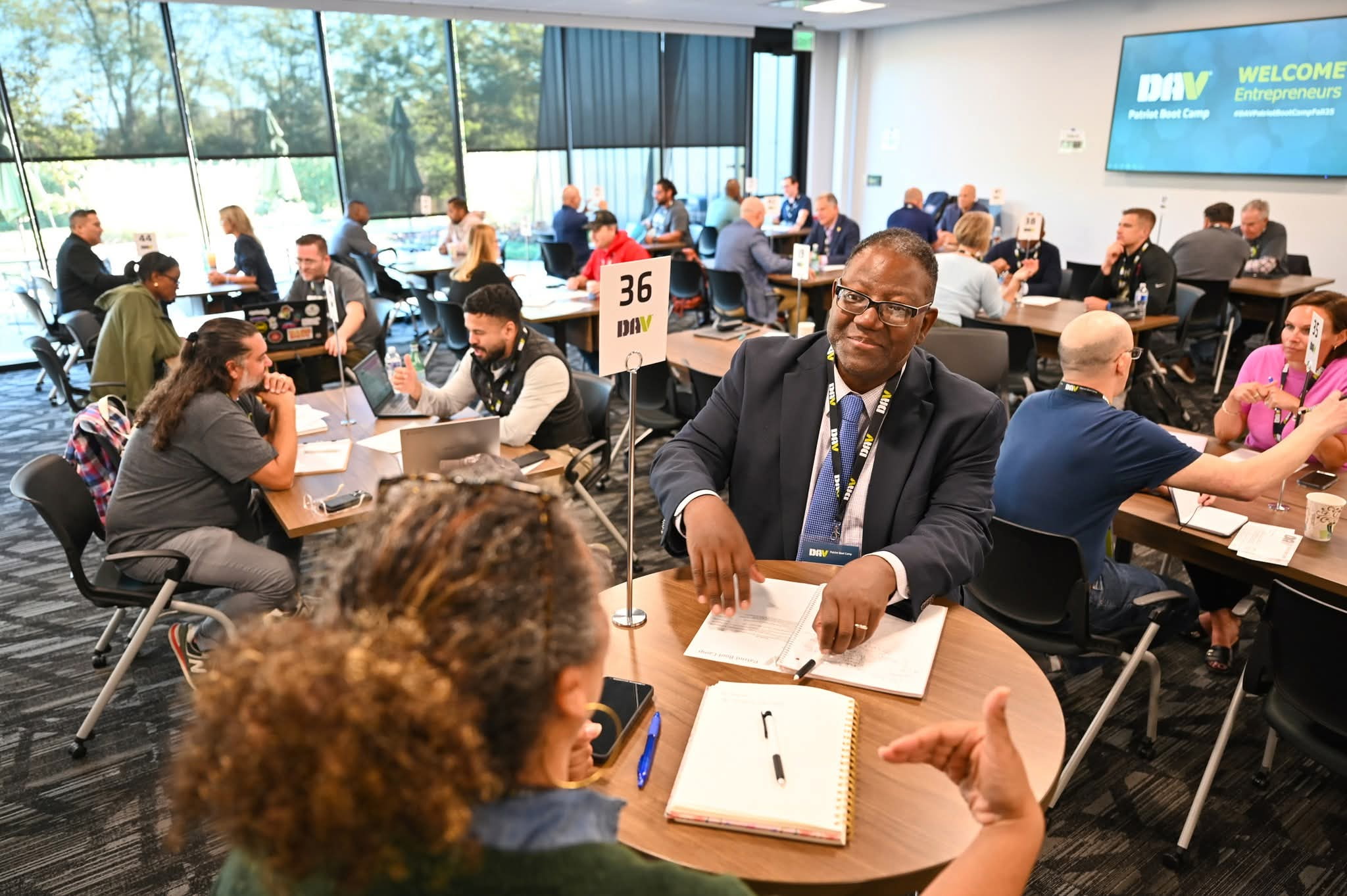 Conference room with people seated at tables, a man in a suit smiling, and a screen displaying a welcome message.