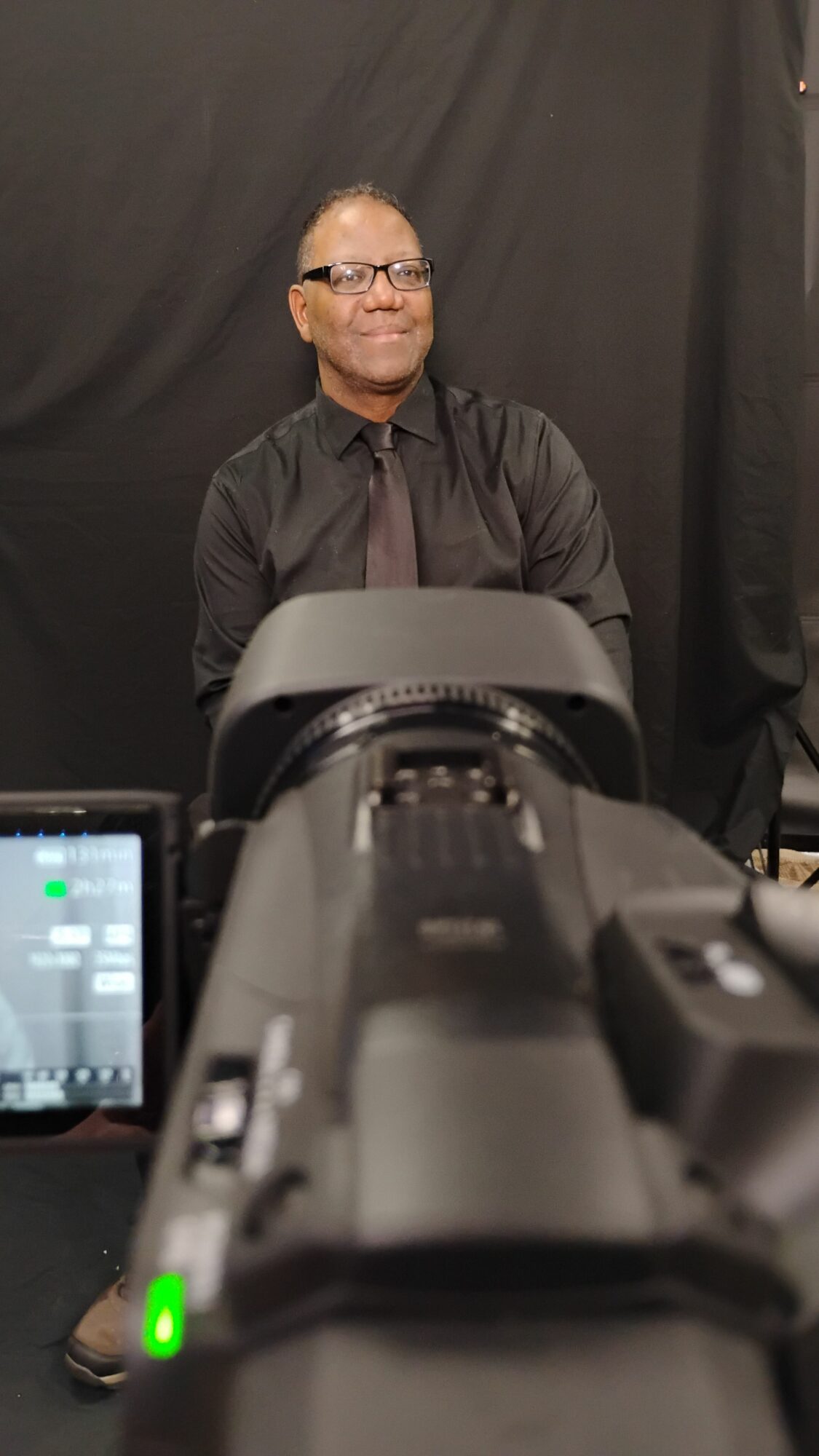 Person with glasses and gray hair, wearing a dark jacket, seen through a camera lens, against a black background.