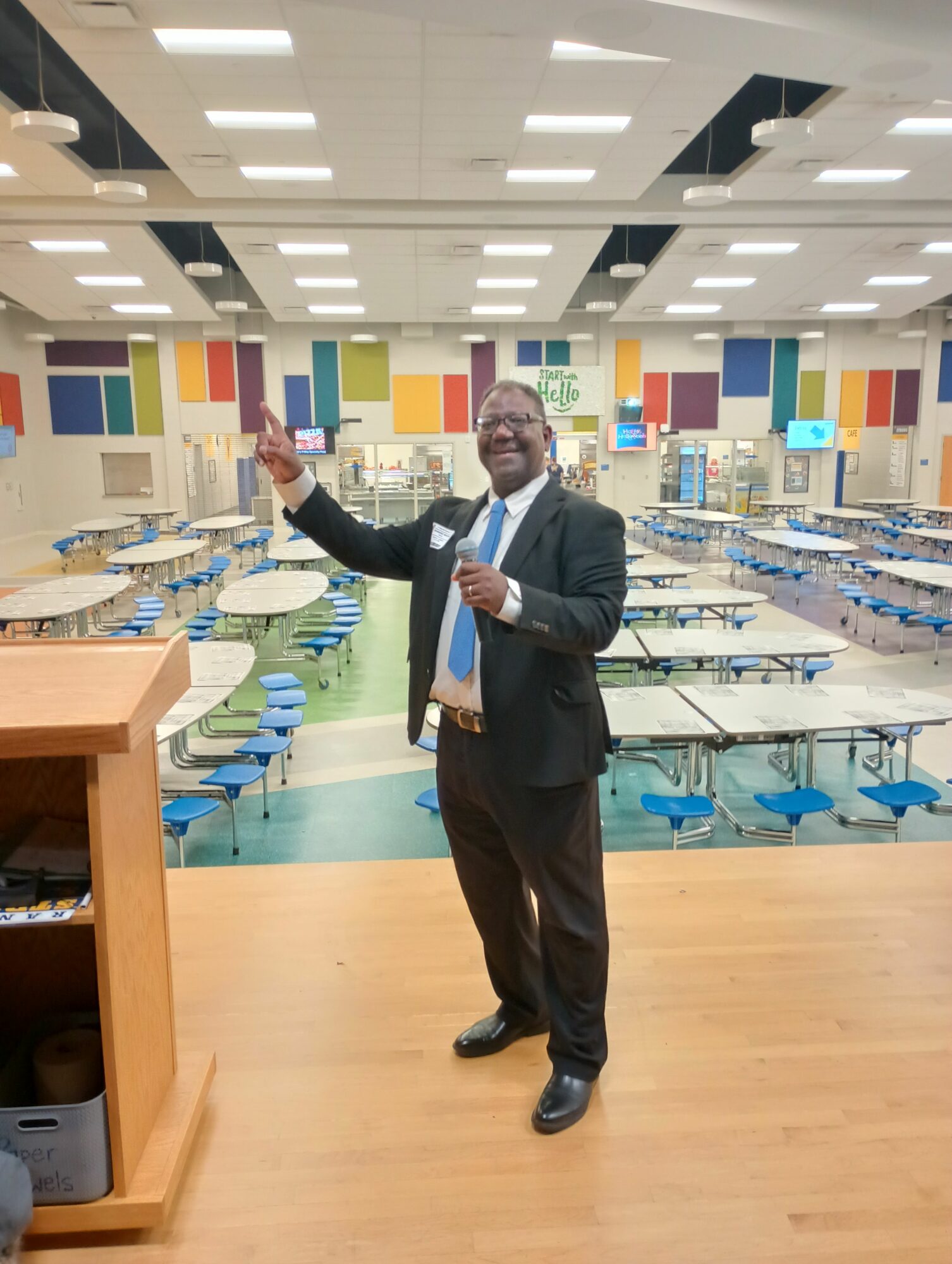 Person in a suit standing in a colorful indoor space, pointing with one hand, with a wooden floor and ceiling lights.