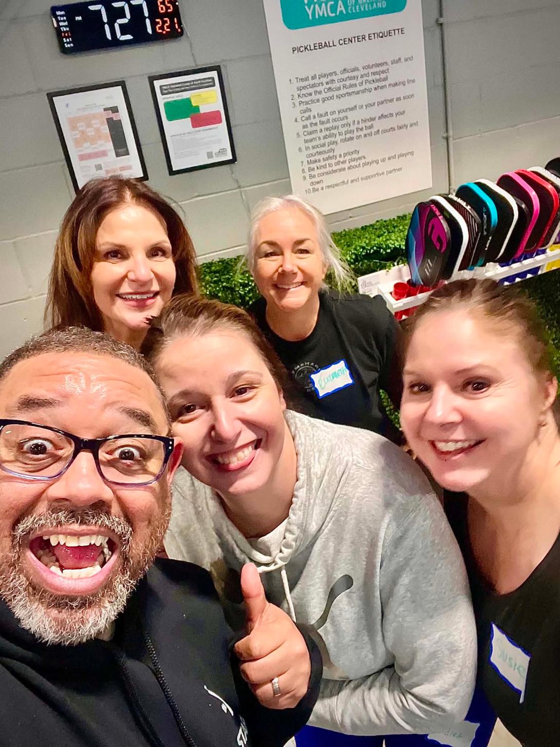 Group of five smiling people taking a selfie indoors, with colorful bags and informational posters in the background.
