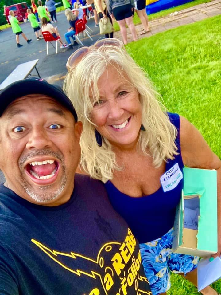 Two smiling people, a man and a woman, outdoors at an event with a crowd and green grass in the background.
