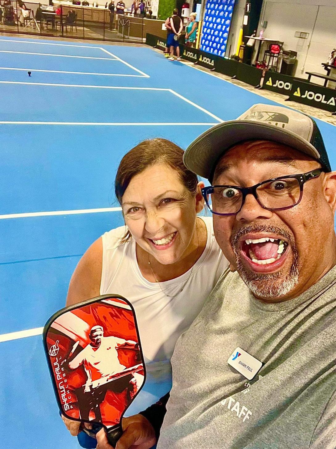 Two people smiling and taking a selfie on a blue tennis court, one holding a paddle, with a background of a sports facility.
