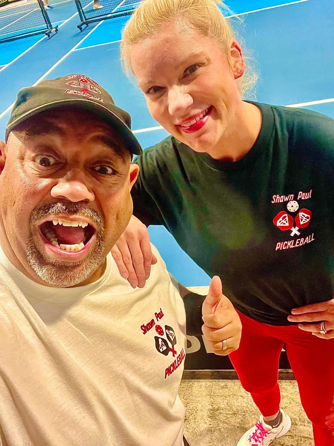 A man and a girl taking a selfie indoors, both smiling, with the girl giving a thumbs-up. The girl wears a black shirt and red pants.