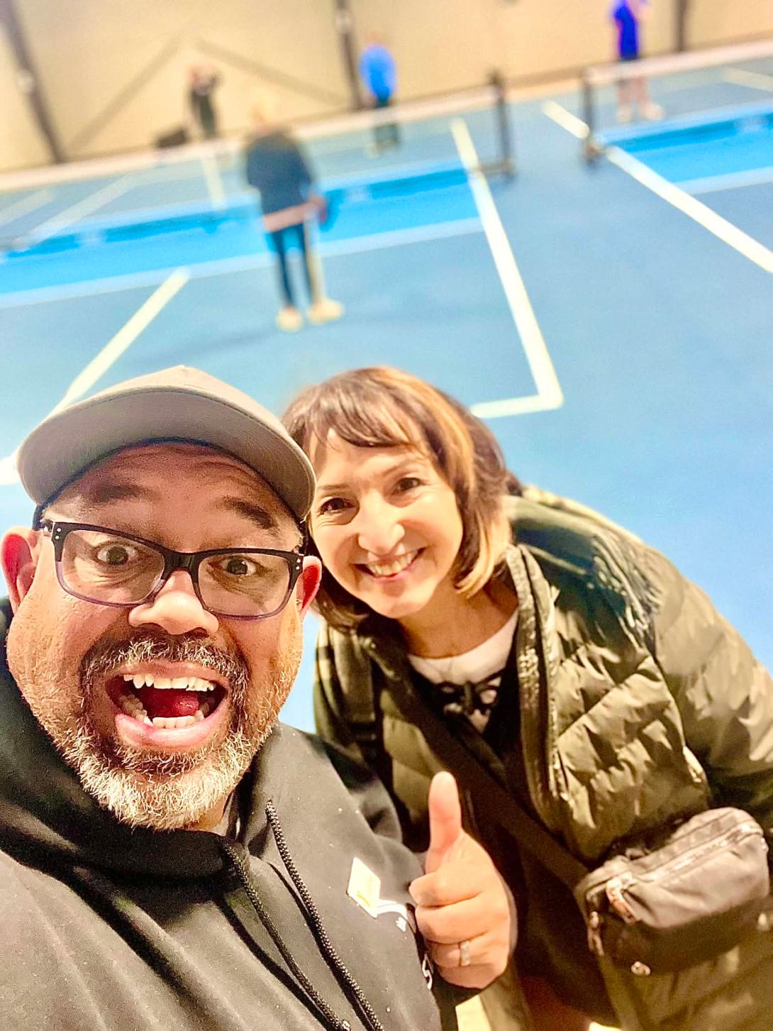Two people smiling and taking a selfie indoors near blue tennis courts, one giving a thumbs-up, with blurred background.