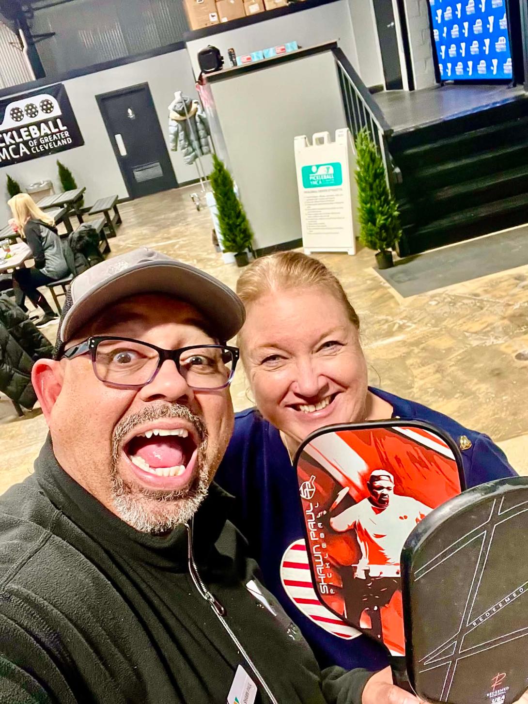 Two people smiling and taking a selfie indoors, with a sports-themed display in the background.