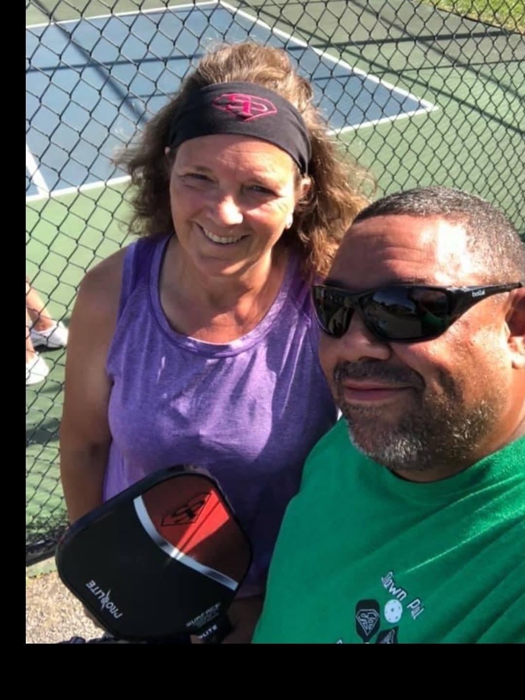 Two people taking a selfie outdoors near a tennis court, smiling, with a chain-link fence in the background.