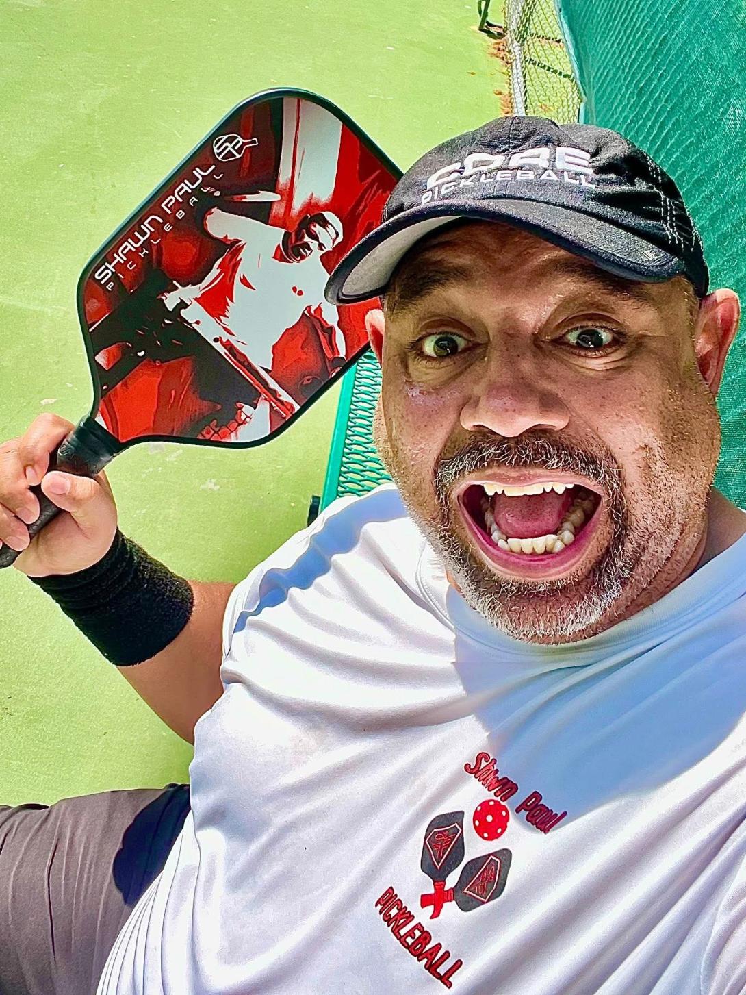Man with a cap smiling and holding a red and black padel racket, wearing a white shirt, in an indoor sports area.