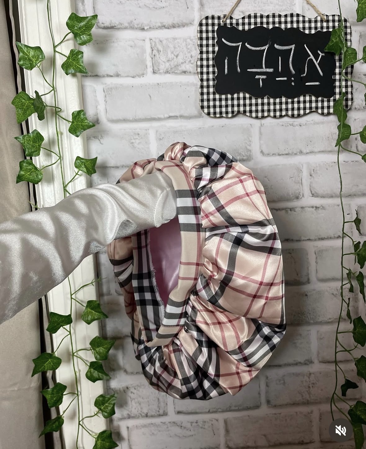 Person's hand holding a plaid fabric bag hanging on a white brick wall with green vines and a black and white sign.