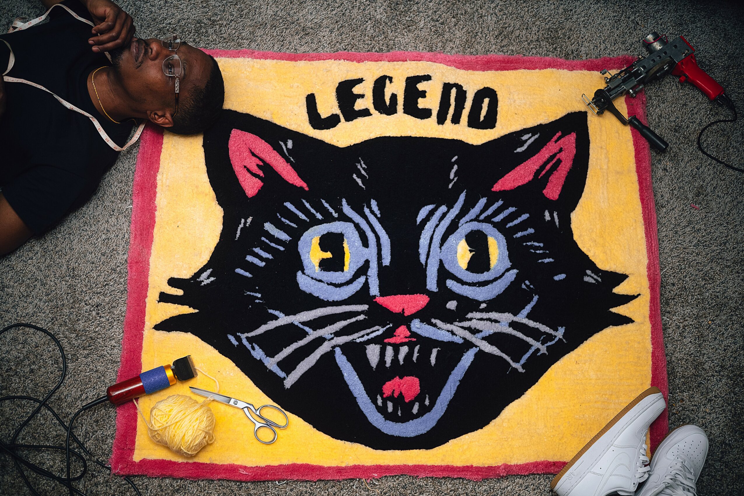 Person lying next to a colorful rug with a large black cat face and the word 'LEGEND' above it, on a gray carpet.
