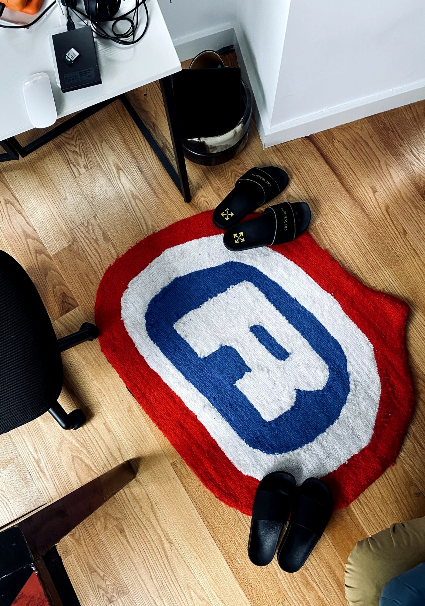 View of a colorful rug with a large letter 'E' in the center, shoes on the rug, and part of a room with wooden floor.