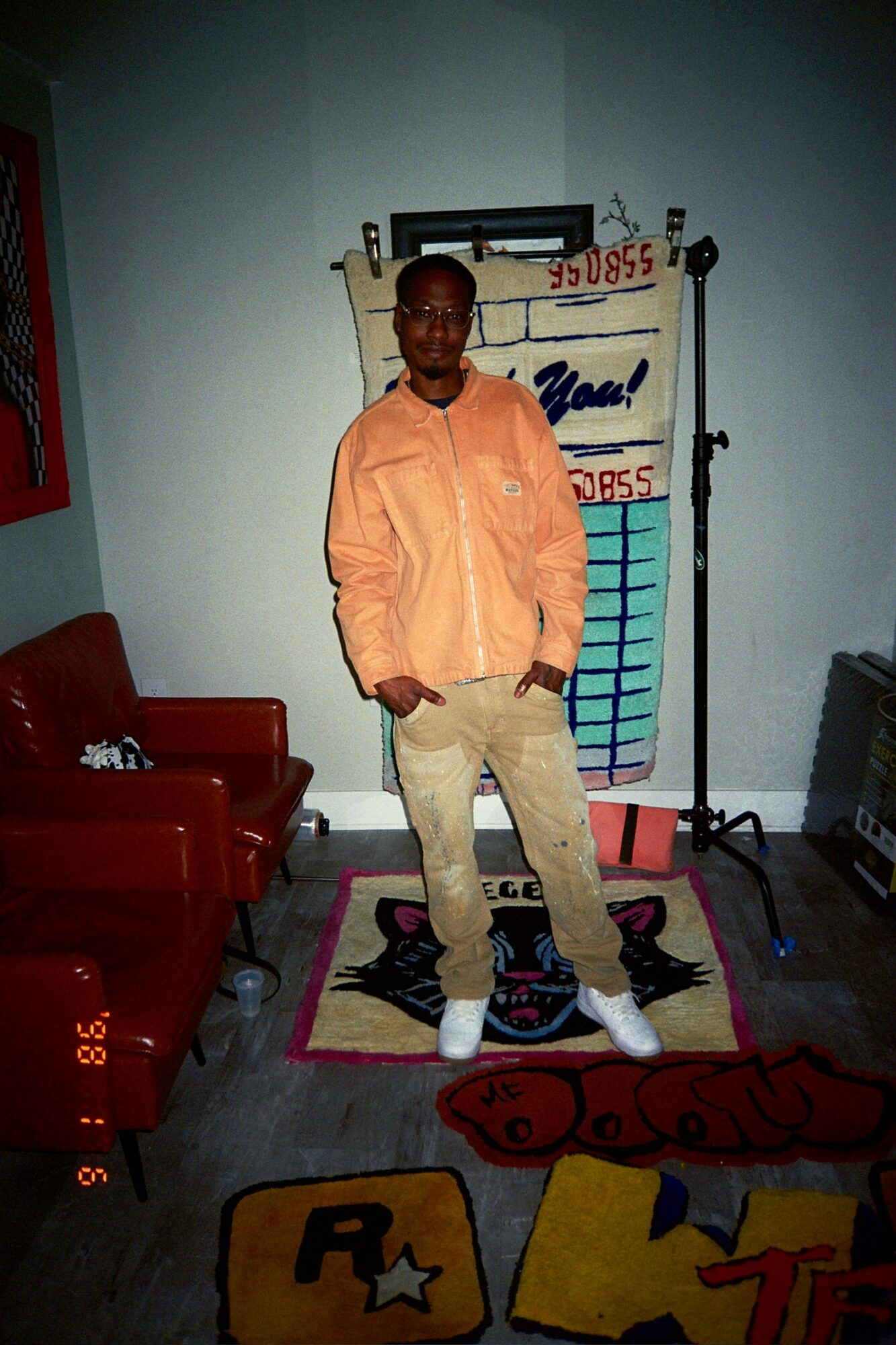 Young man standing on a colorful rug in a room with chairs and a display board behind him.