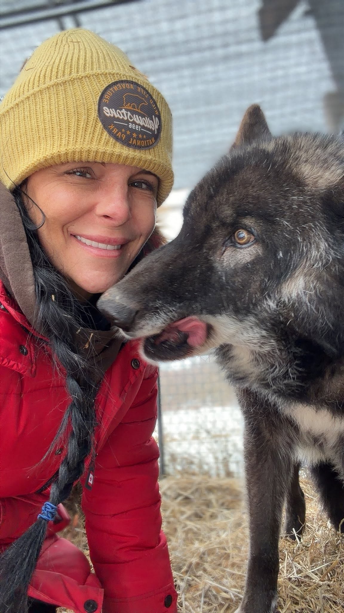 Person wearing a yellow beanie and red jacket smiling with a black and gray dog outdoors.