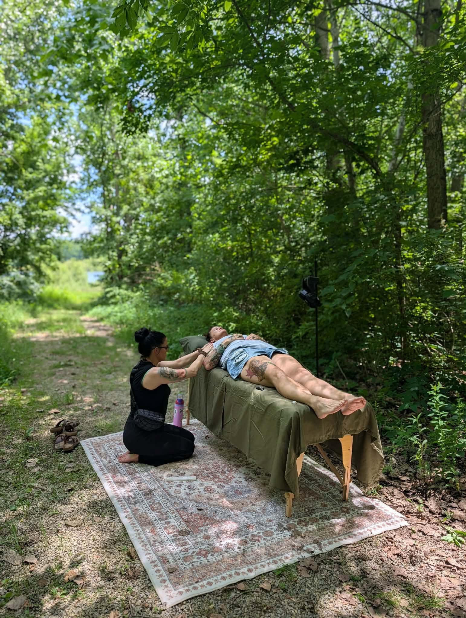 Person lying on a table outdoors receiving a massage from another person, surrounded by trees and greenery, on a rug.