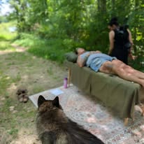 Person lying on a table outdoors with a dog nearby, and a woman standing in a wooded area.