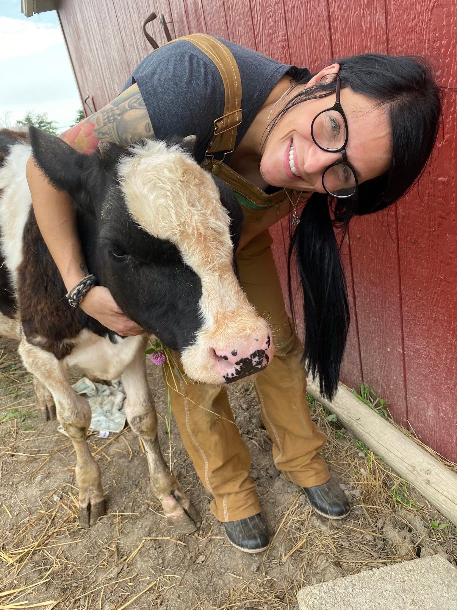 Person with glasses and tattoos hugging a calf outdoors, smiling and leaning towards it, with a red wooden wall in the background.