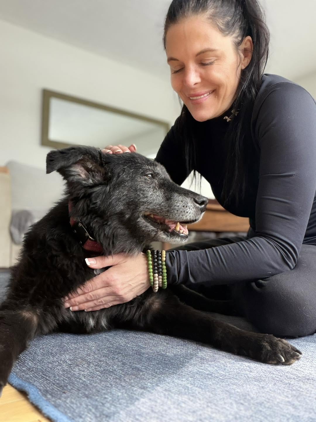 Woman smiling and petting a black dog lying on a carpeted floor indoors.
