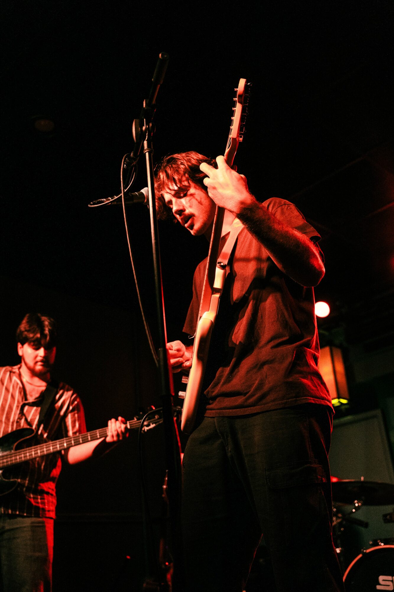 Musician playing guitar on stage with another musician in background, dark setting, stage lighting, focused on performer.