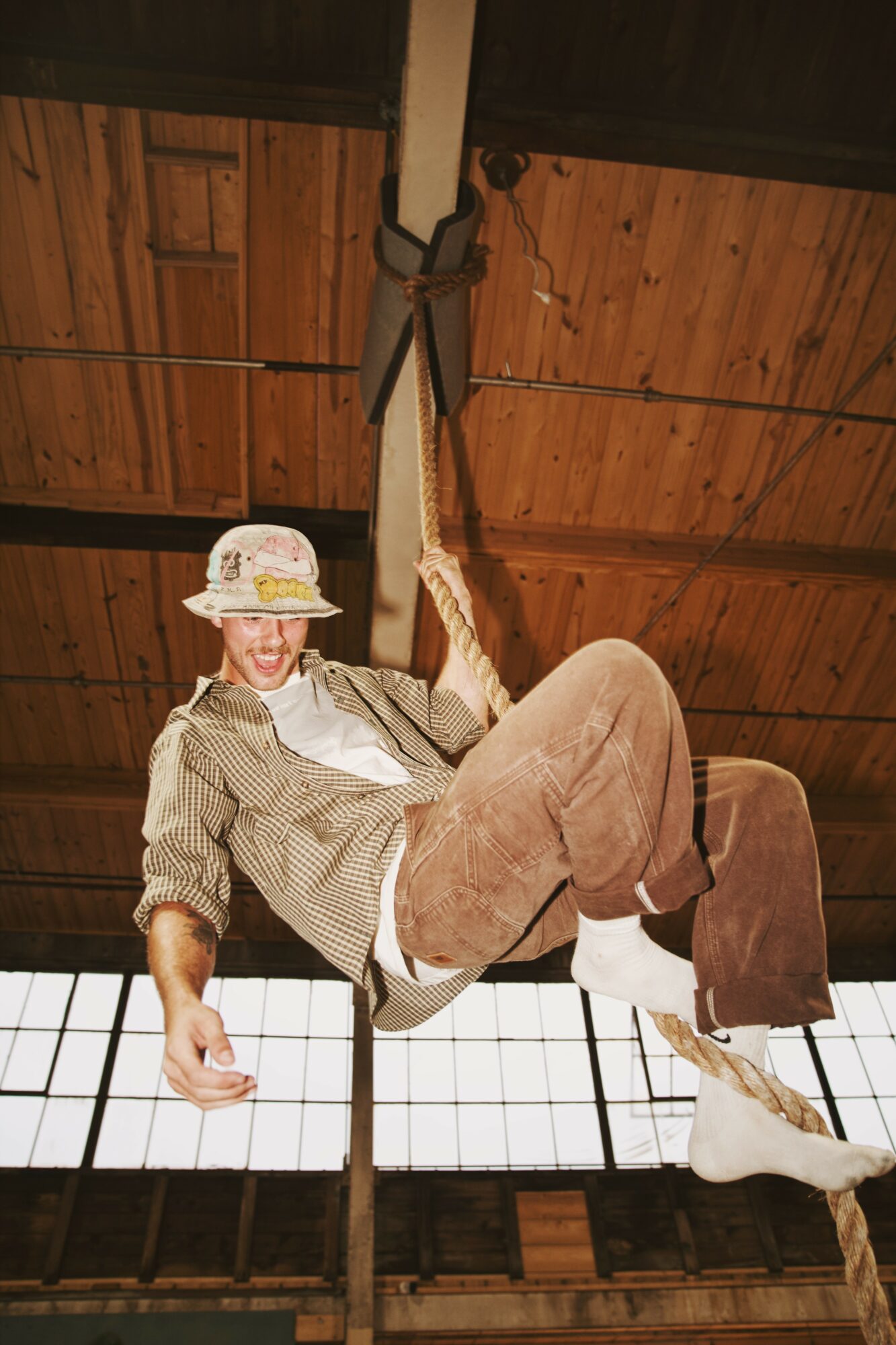 Person hanging from a rope inside a wooden structure, smiling, wearing a hat, shirt, and pants.