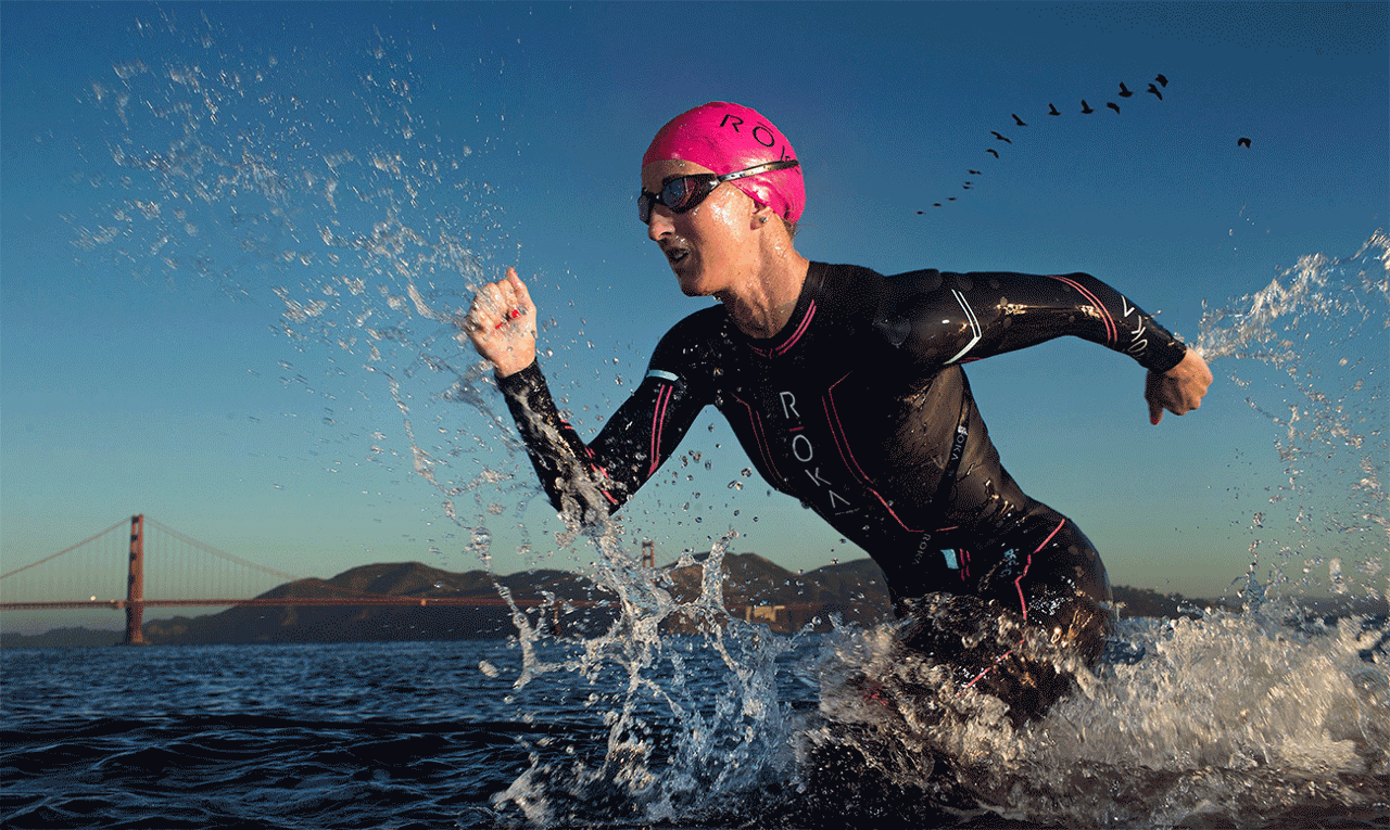 Person running through water wearing a pink swim cap and black wetsuit, with a bridge and hills in background.