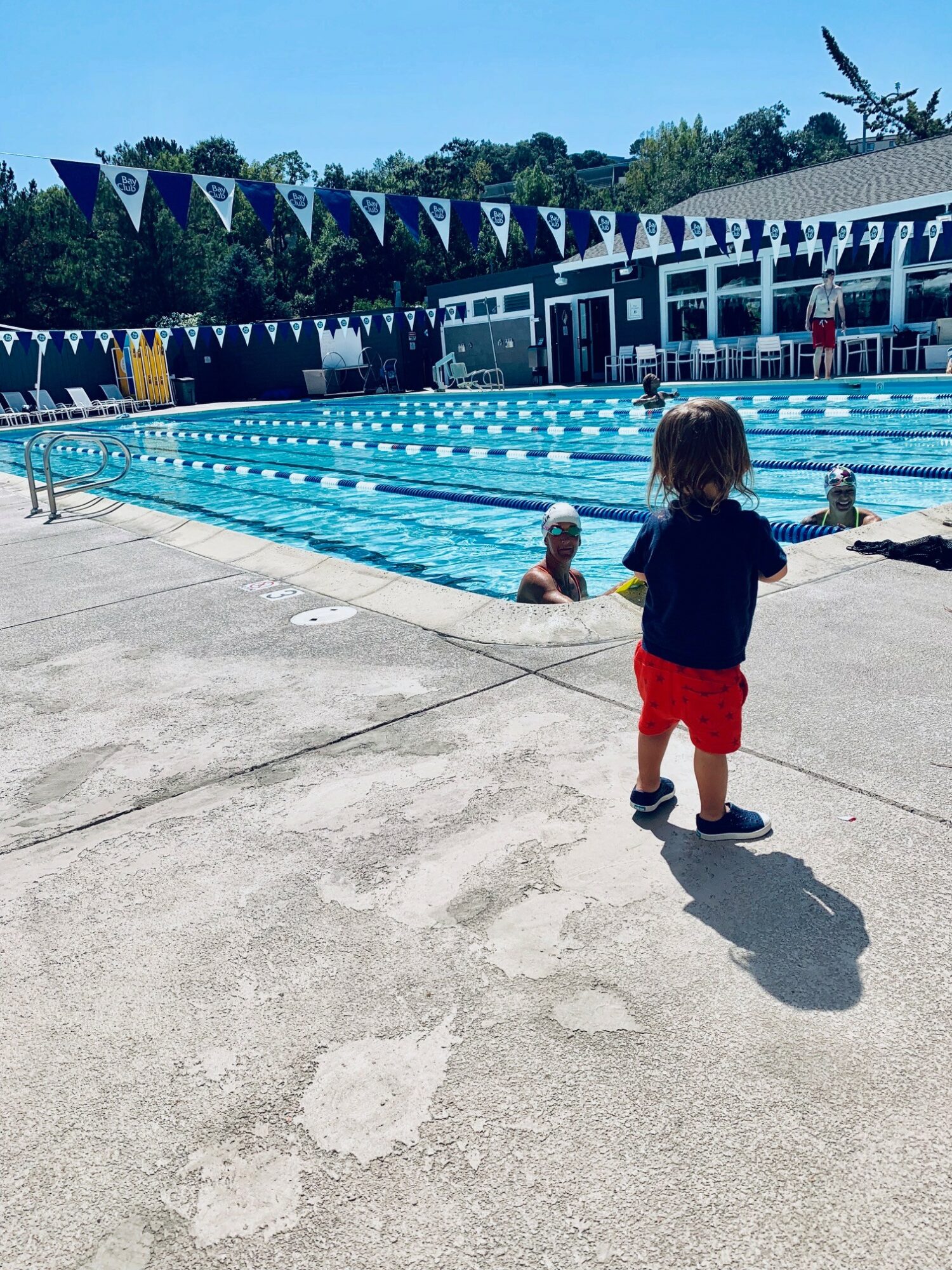 Child in red shorts and dark shirt walking near a swimming pool with people swimming, pool flags, and a building in the background.