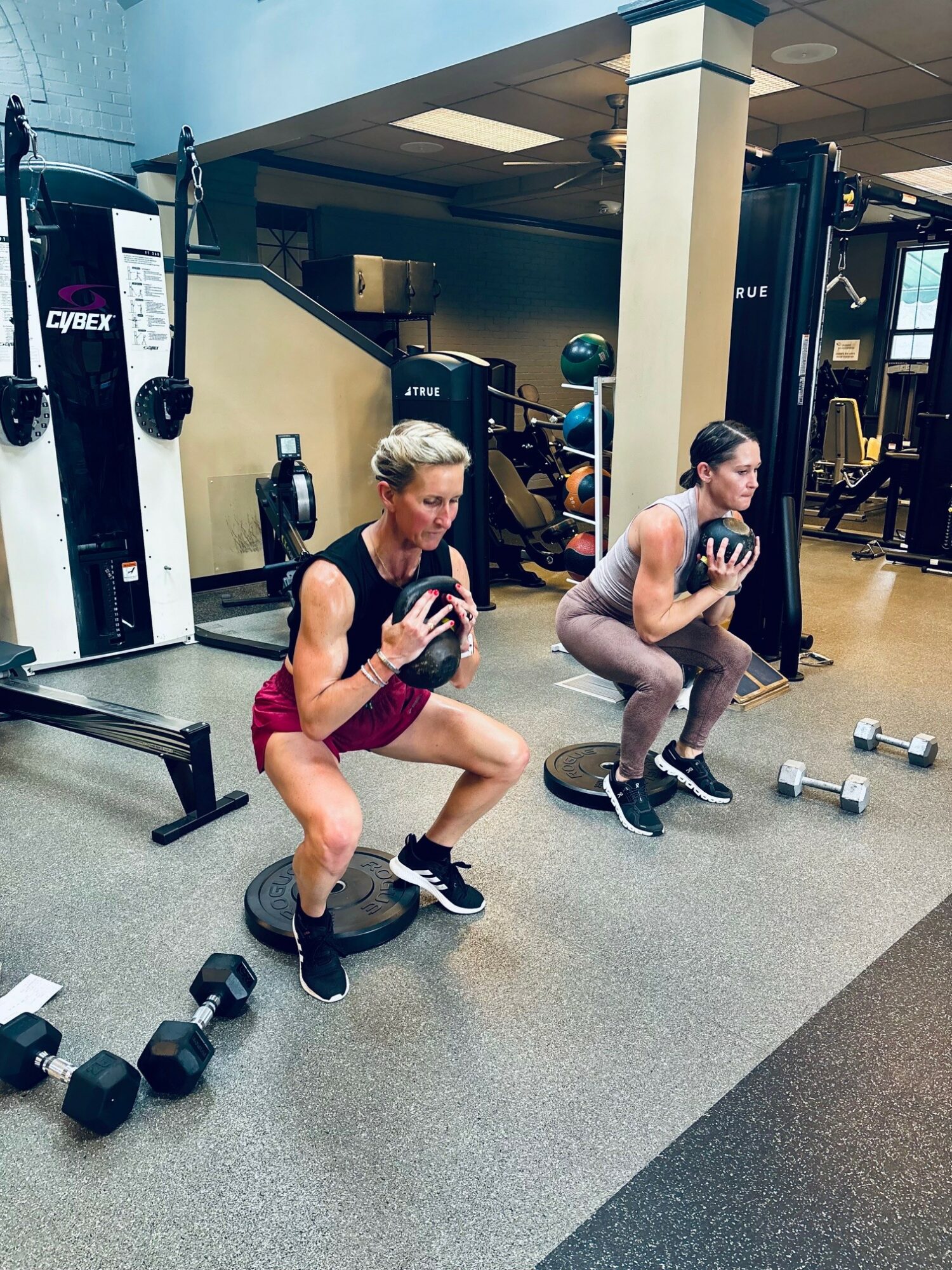 Two women perform squats with weights in a gym, surrounded by exercise equipment and dumbbells.
