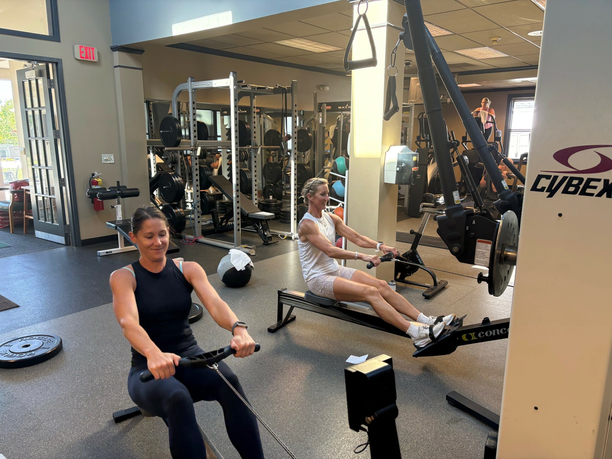 Two women exercising on rowing machines in a gym with workout equipment in the background.