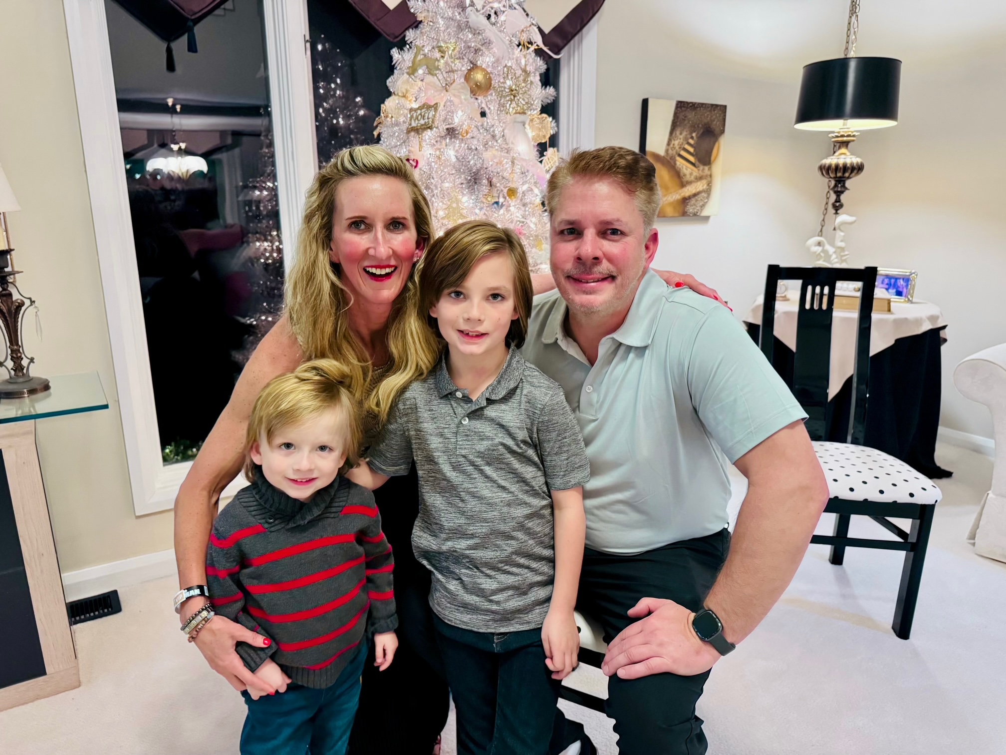 Family of four smiling indoors near a decorated Christmas tree, with a woman, two children, and a man, in a living room.