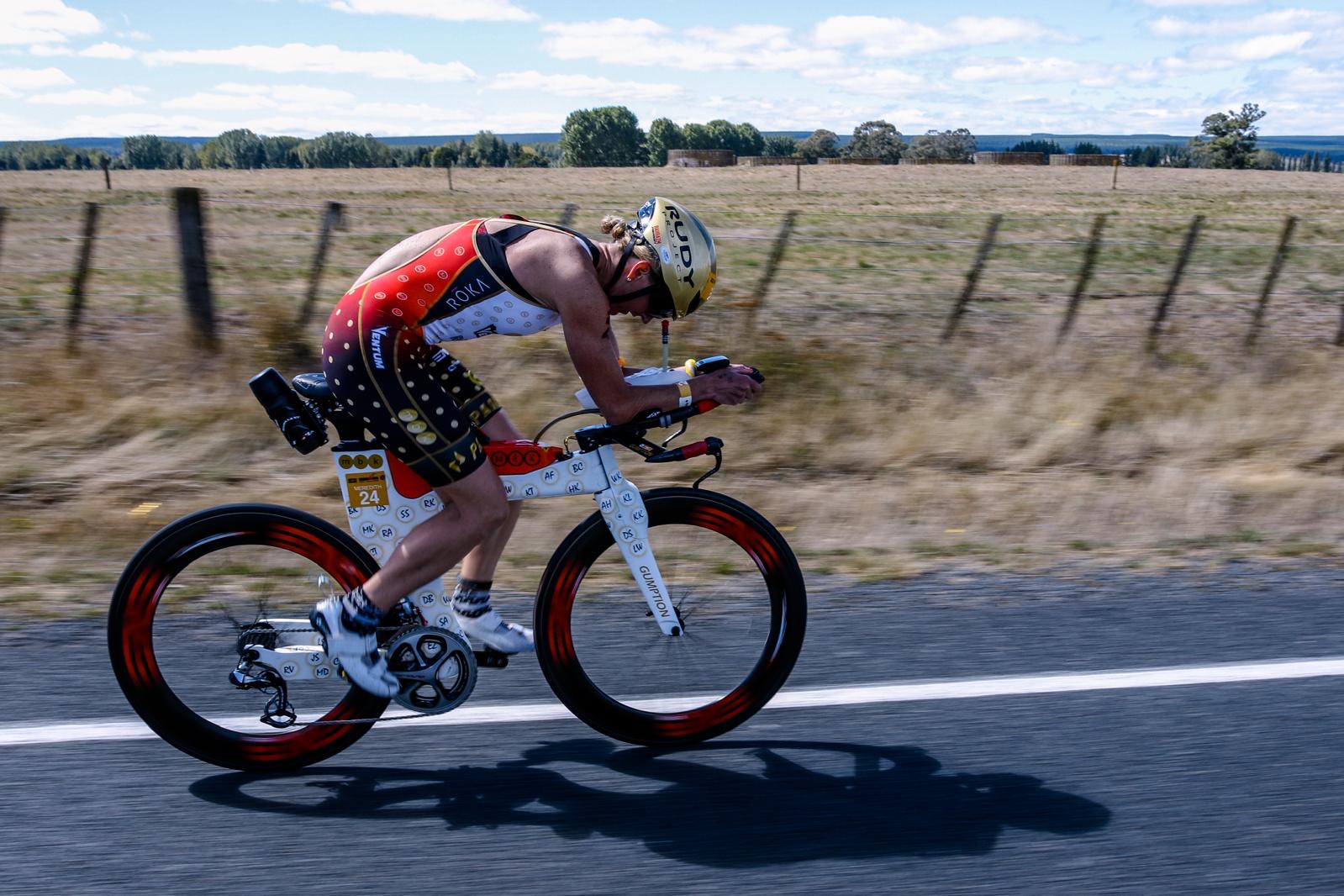 Cyclist wearing a helmet and athletic clothing rides a mountain bike on a road with a grassy field and fence in background.