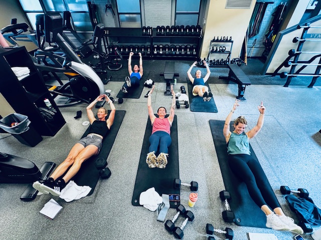 Five women lying on exercise mats with arms raised in a gym, surrounded by workout equipment and weights.