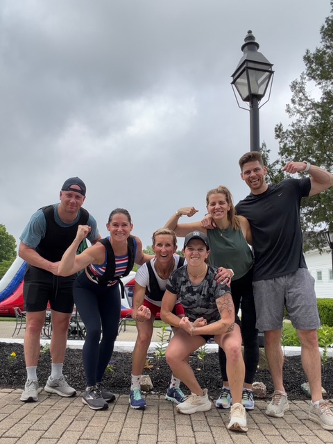 Group of six people outdoors, smiling, posing with flexed arms, near a street lamp and trees, cloudy sky in background.