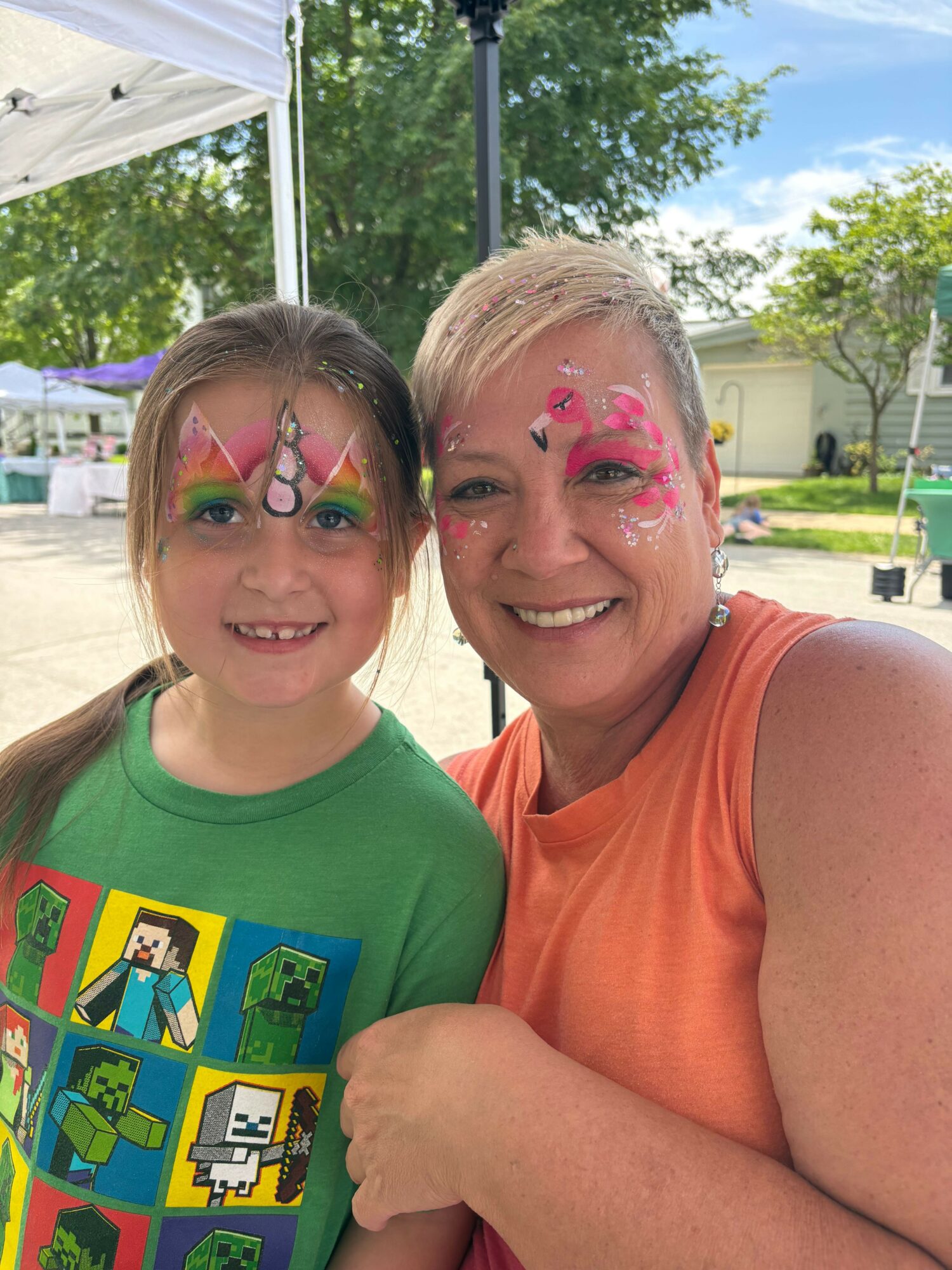 Two smiling children with colorful face paint, outdoors on a sunny day, one wearing an orange shirt, the other a green shirt.