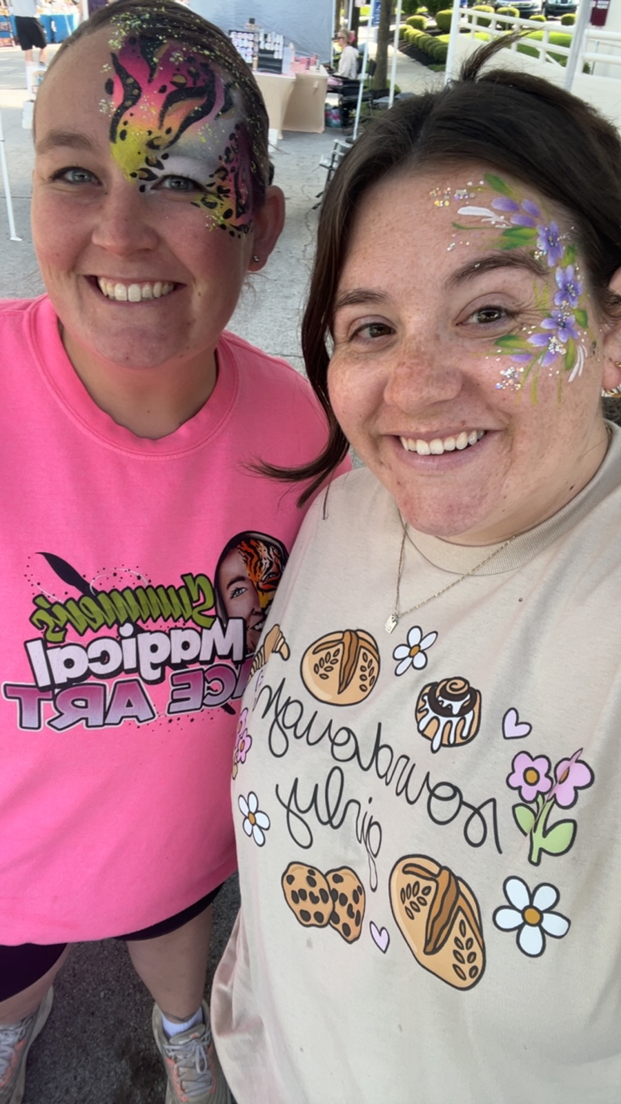 Two women smiling, one with colorful face paint, wearing casual clothing, outdoors with trees and structures in background.