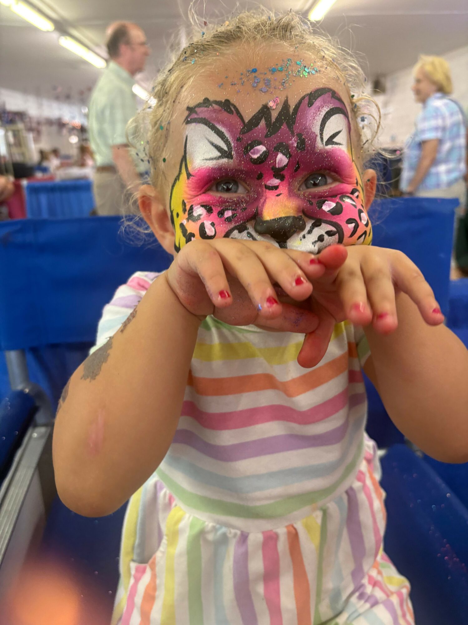 Child with face painted as a pink and black tiger, wearing a striped dress, holding a yellow object, in a crowded setting.