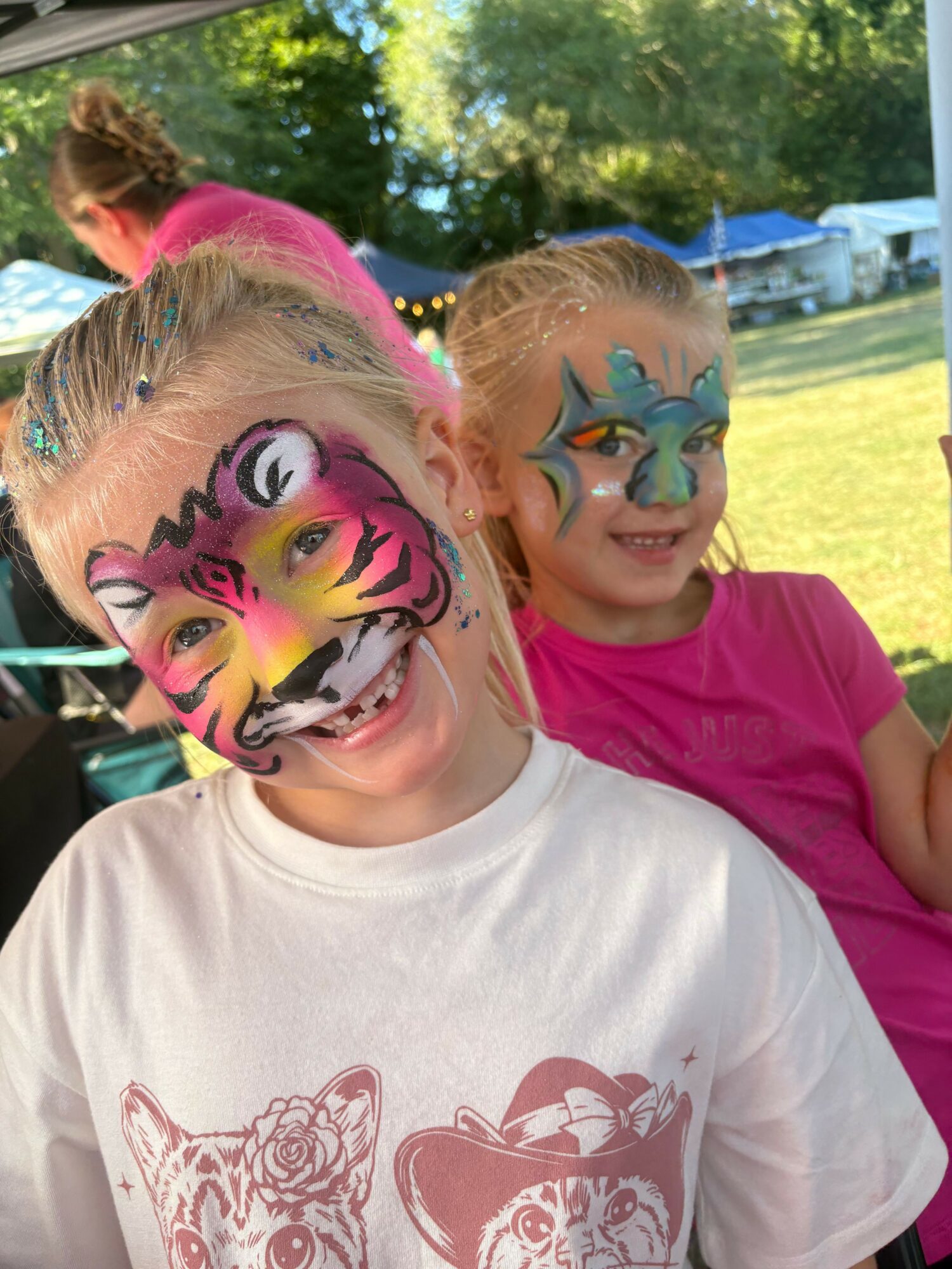 Two children with colorful face paint, outdoors with trees and grass in the background.