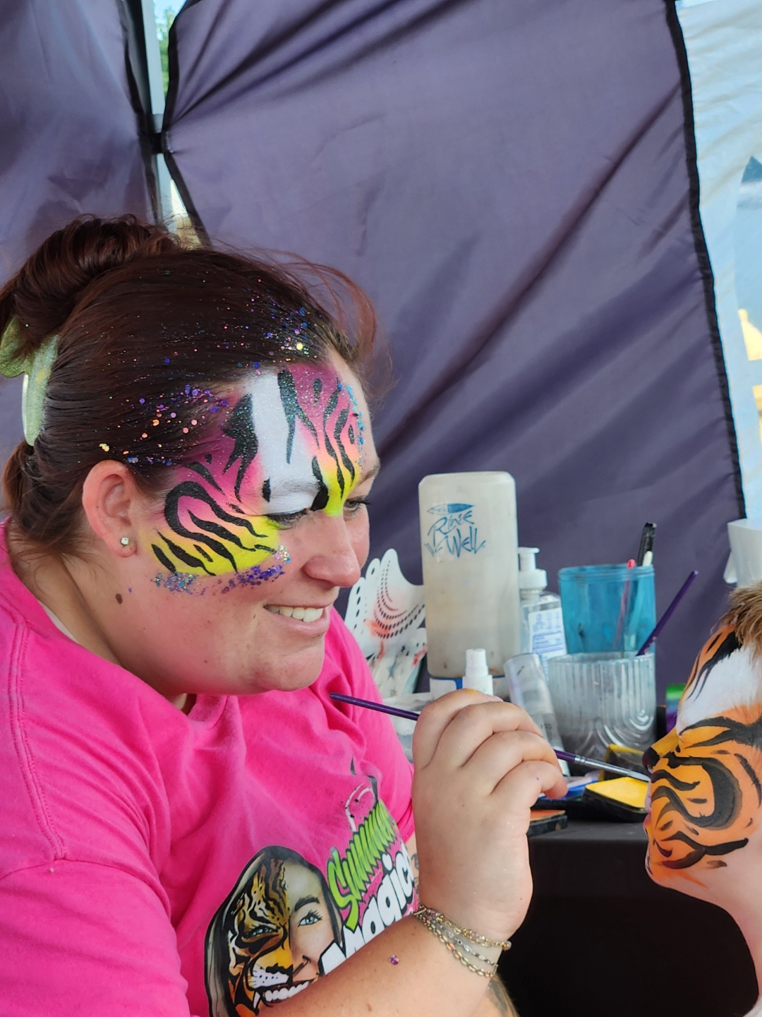 Woman with colorful tiger face paint, smiling, holding a paintbrush, inside a tent with art supplies visible.