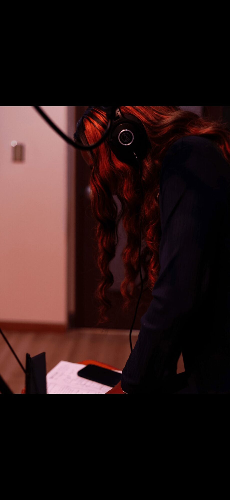 Person with long, wavy hair wearing dark clothing, standing indoors near a table with a phone, face obscured by headphones.