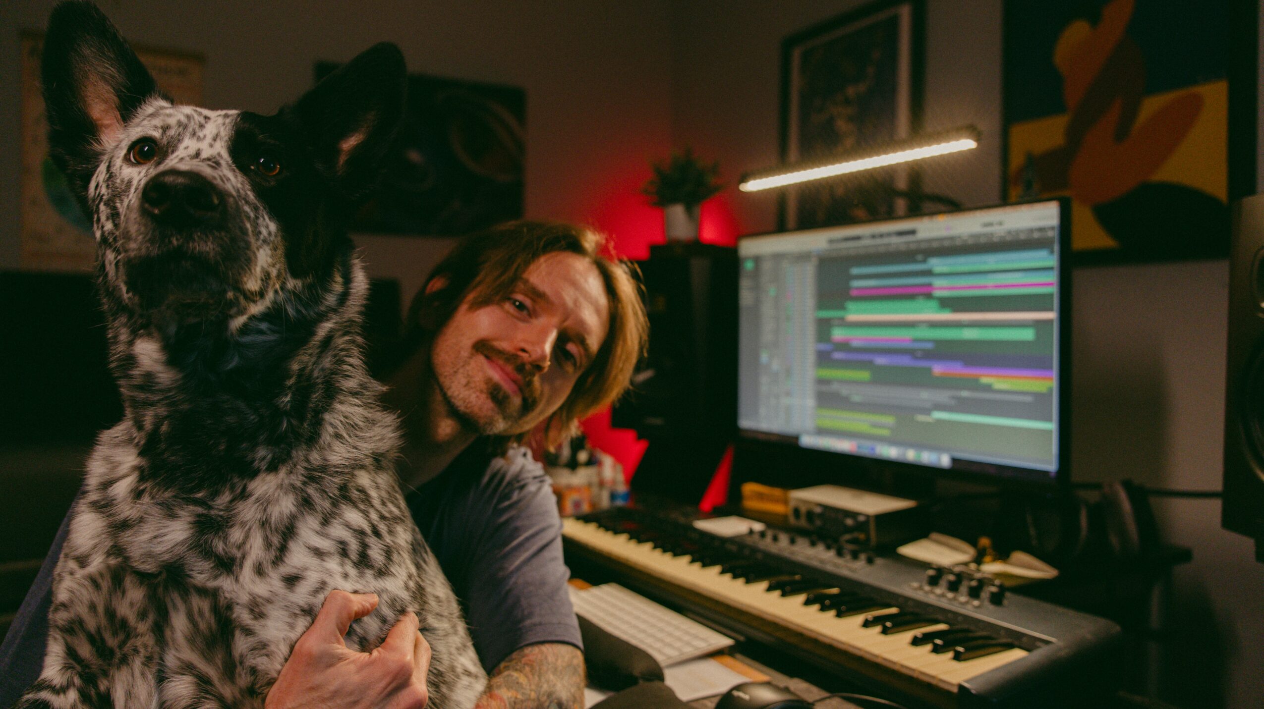 Person smiling with a dog in a music studio with a computer and keyboard, colorful artwork on the wall.