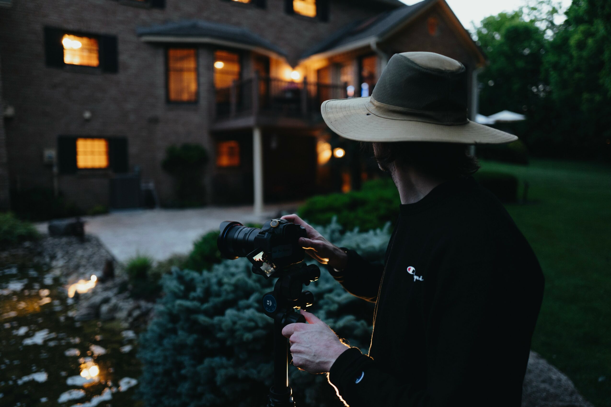 Person wearing a wide-brimmed hat holding a camera outdoors near a house at dusk.