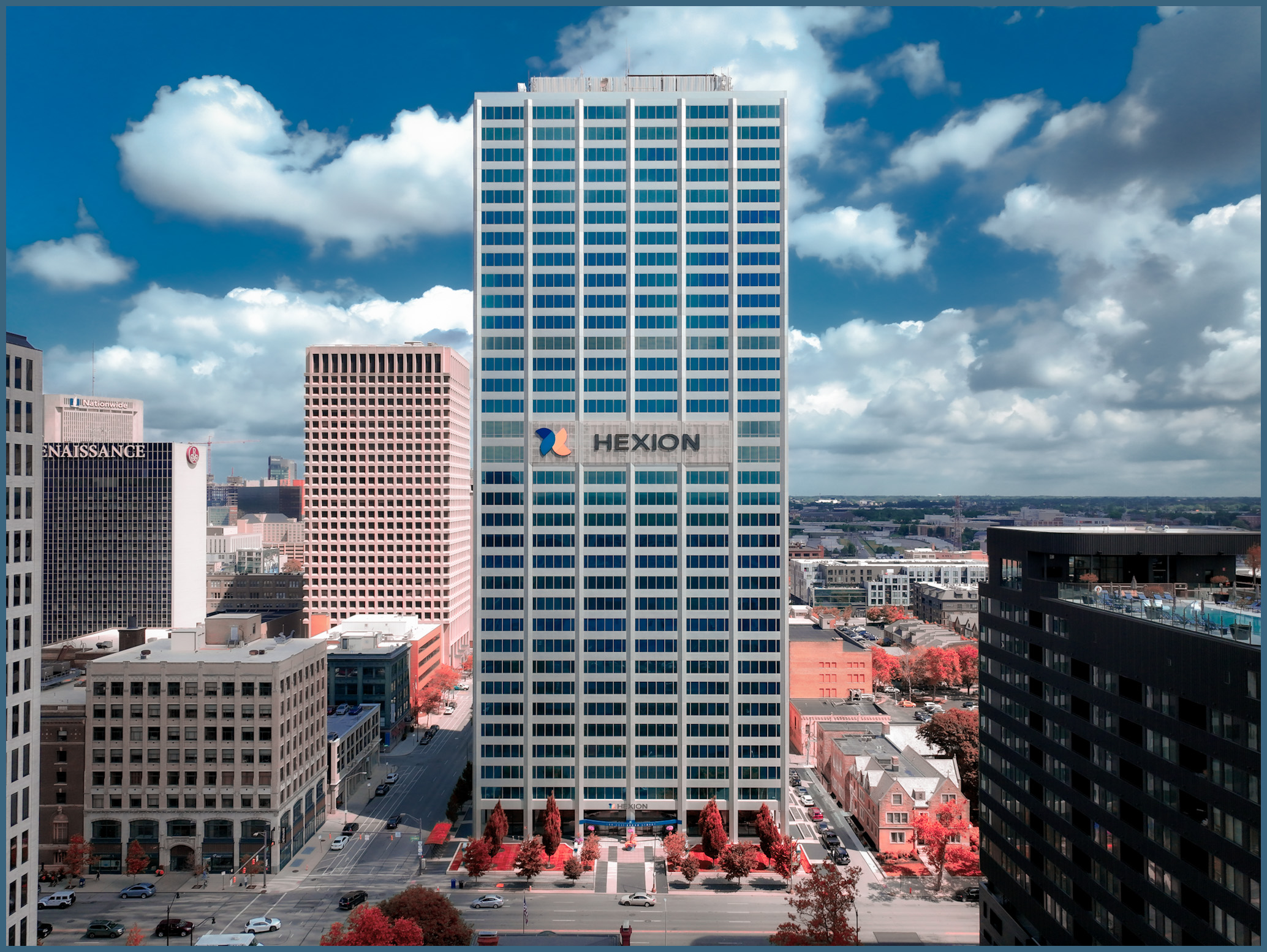 Tall white and blue office building with the Hexion logo, surrounded by other buildings under a partly cloudy sky.