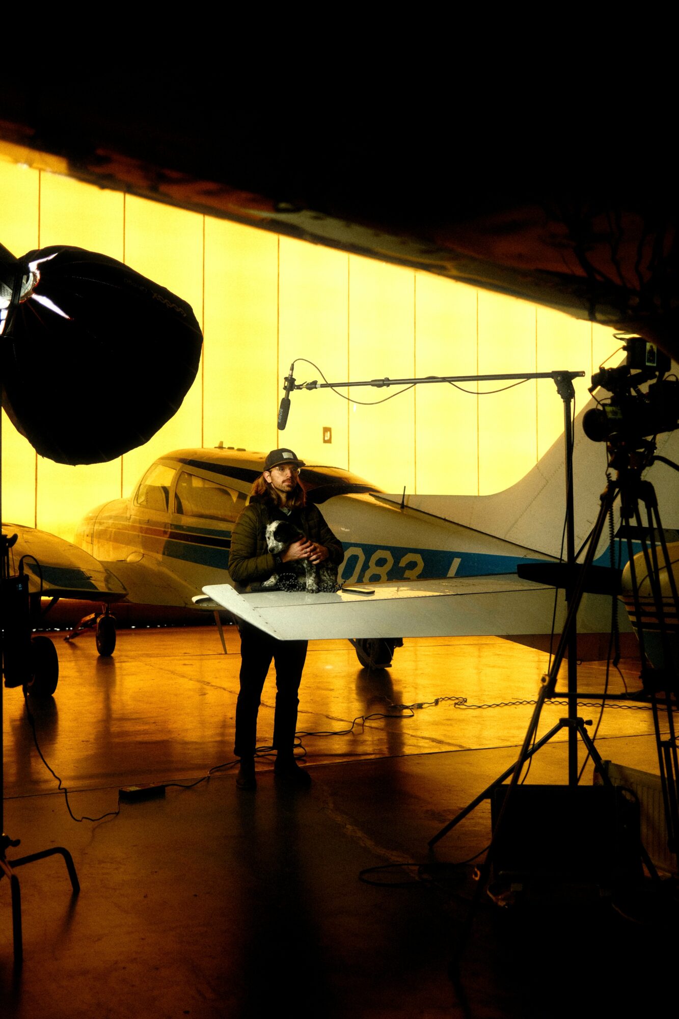 Person working in a studio with cameras, lights, and a yellow background, holding a large sheet of paper or board.