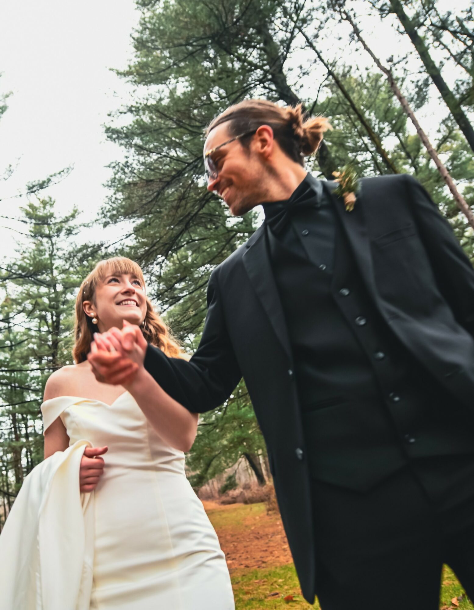 A man and woman shake hands outdoors among trees, smiling at each other, dressed formally, woman in a white dress, man in a black suit.