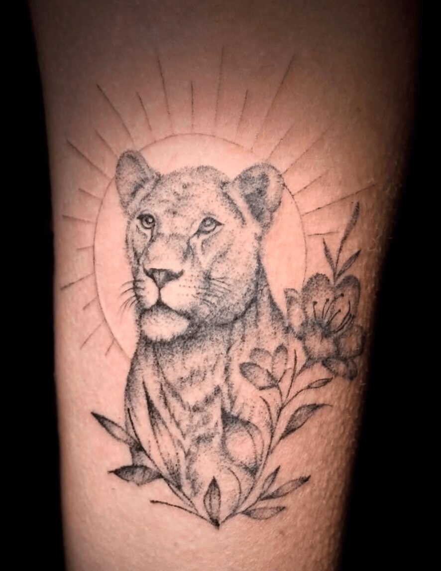 A tattoo of a lioness's head surrounded by flowers and radiating lines, in black ink on skin.