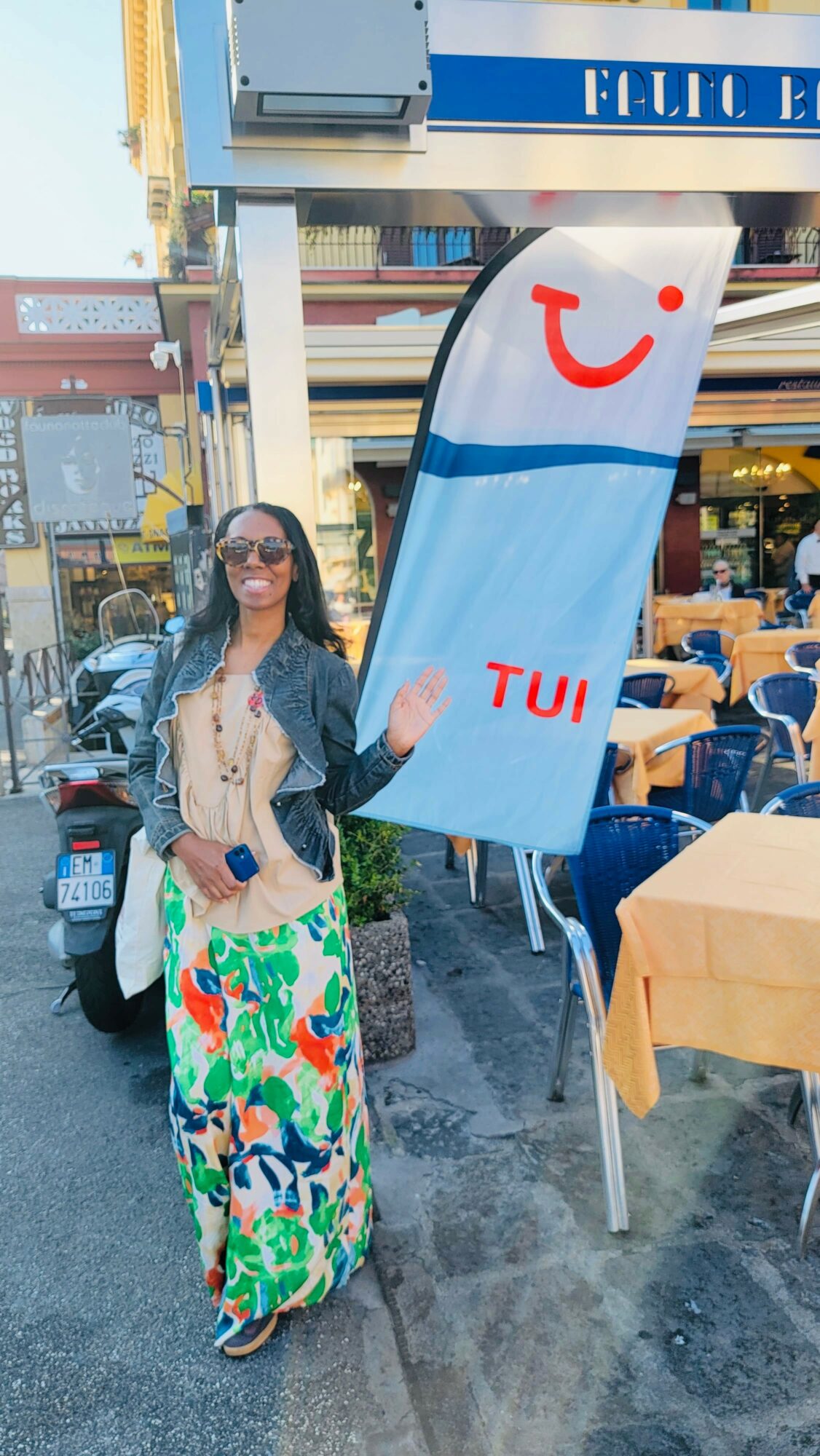 Woman standing on a busy street with a large TUI sign, wearing sunglasses and a colorful skirt, next to outdoor tables.