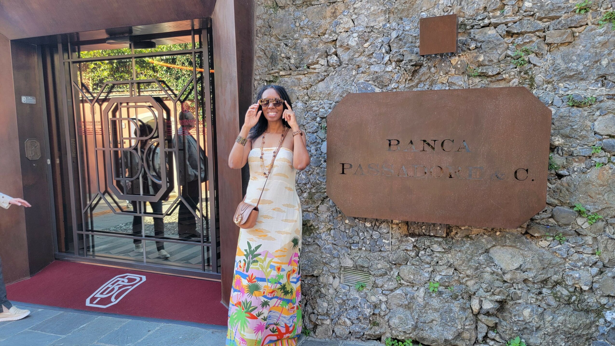 Woman in a colorful dress and sunglasses standing outside near a stone wall and a metal gate.