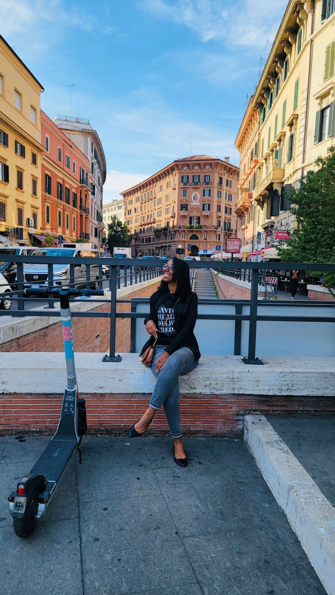 Person sitting on a ledge in a city street with colorful buildings and a blue sky.