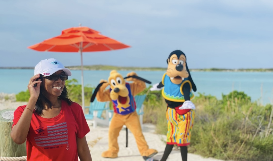 Person in red shirt and white cap smiling at the beach with two costumed characters, a dog and a woman, near water and greenery