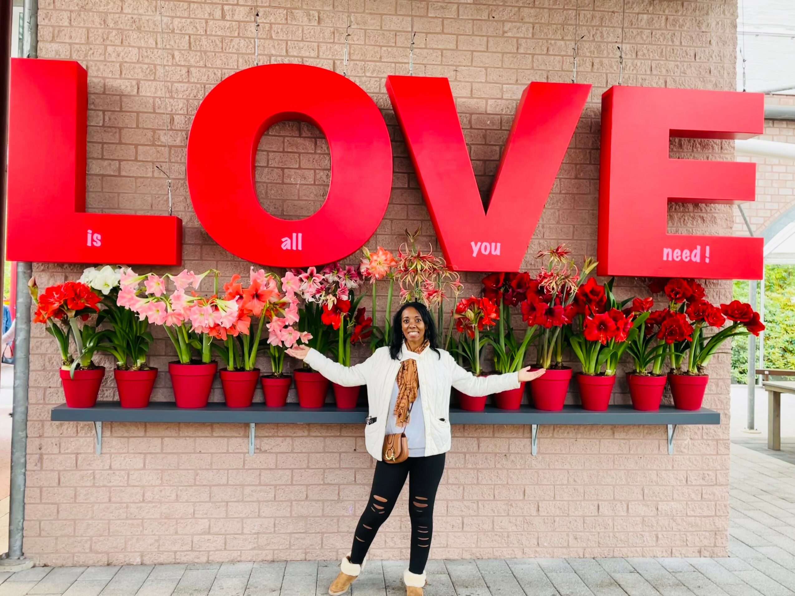 Person standing in front of large red letters spelling LOVE, with pink and red flowers in pots below, on a brick wall.