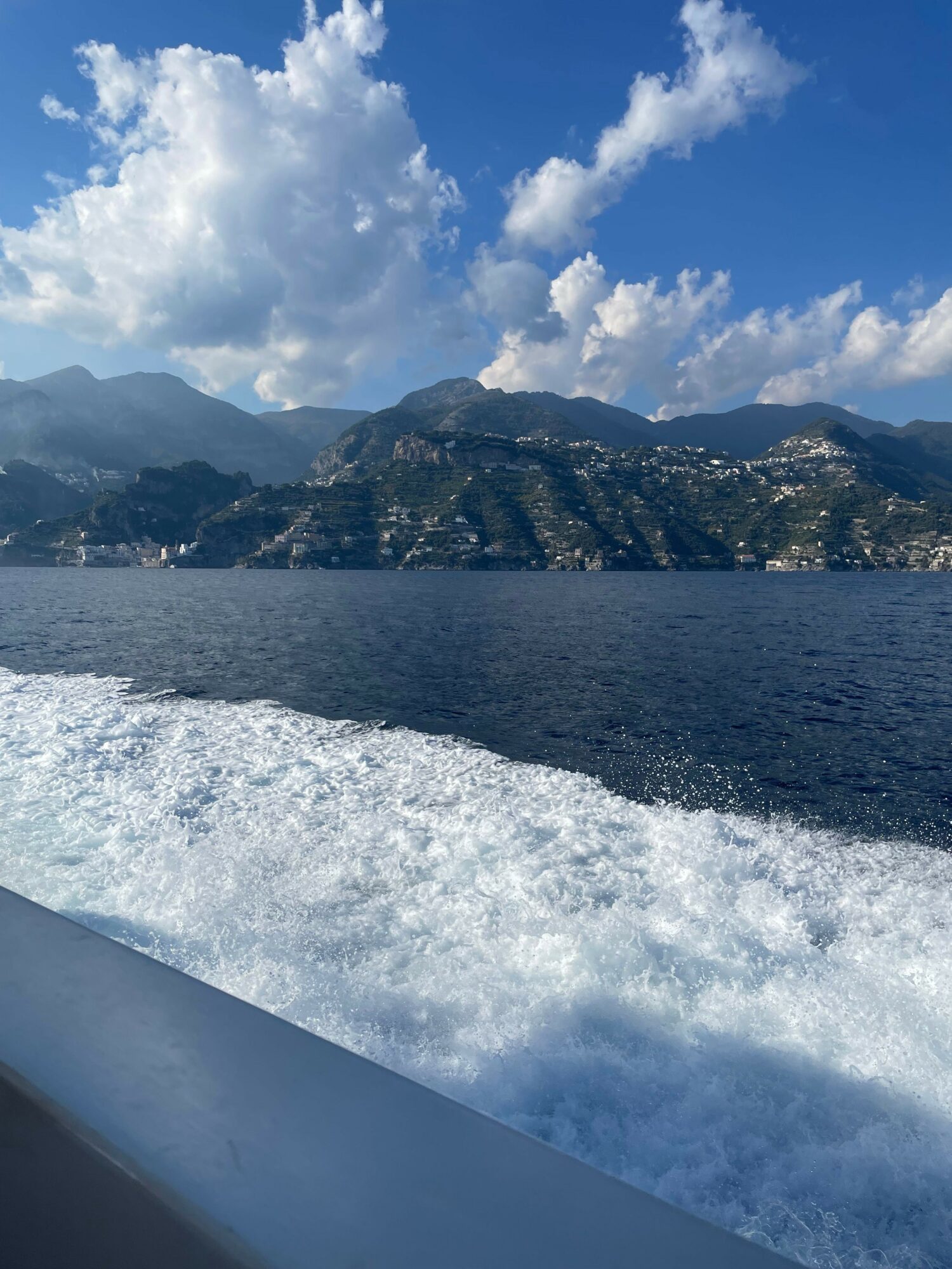 View of a mountain landscape with water and sky, seen from a boat moving through water, with clouds and mountains in the background.
