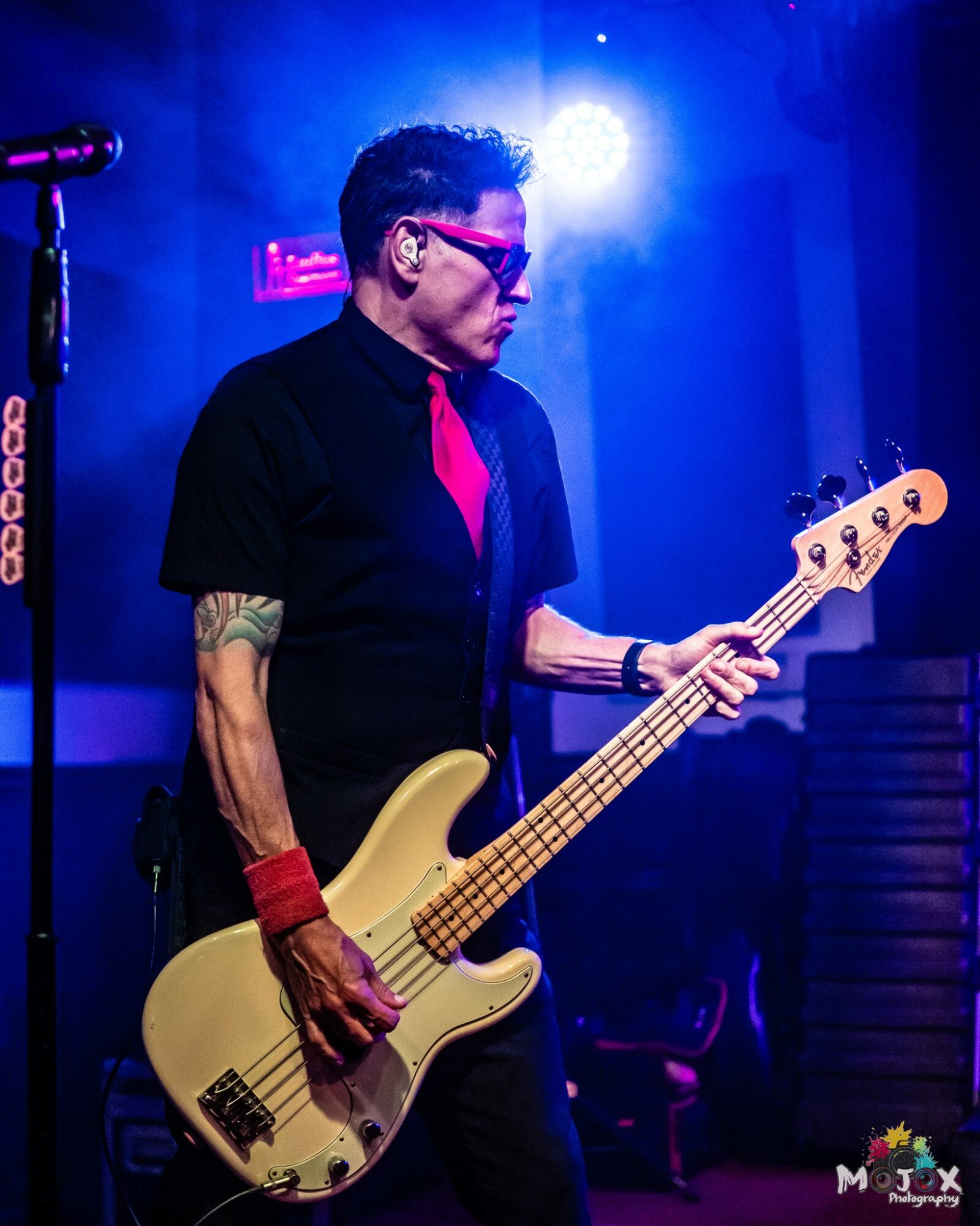Person playing bass guitar on stage with blue lighting, wearing sunglasses, black shirt, and red accessories.